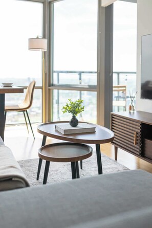 Ein modernes Wohnzimmer mit einer Couch, einem Couchtisch, einem kleinen Beistelltisch und einem Bücherregal. Ein großes Fenster bietet Blick auf die Stadtlandschaft.