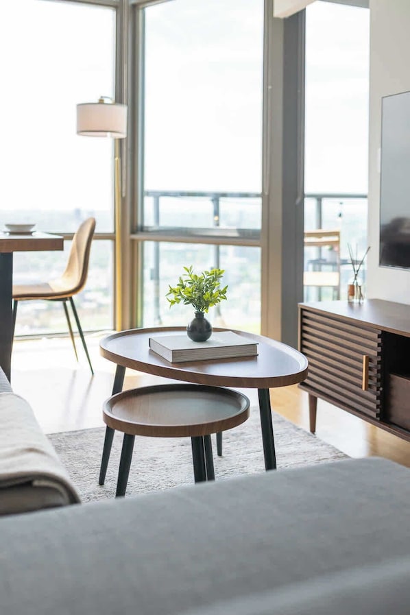 A modern living room with a sofa, a coffee table, a small side table, and a bookshelf. There is a large window providing a view of the cityscape.