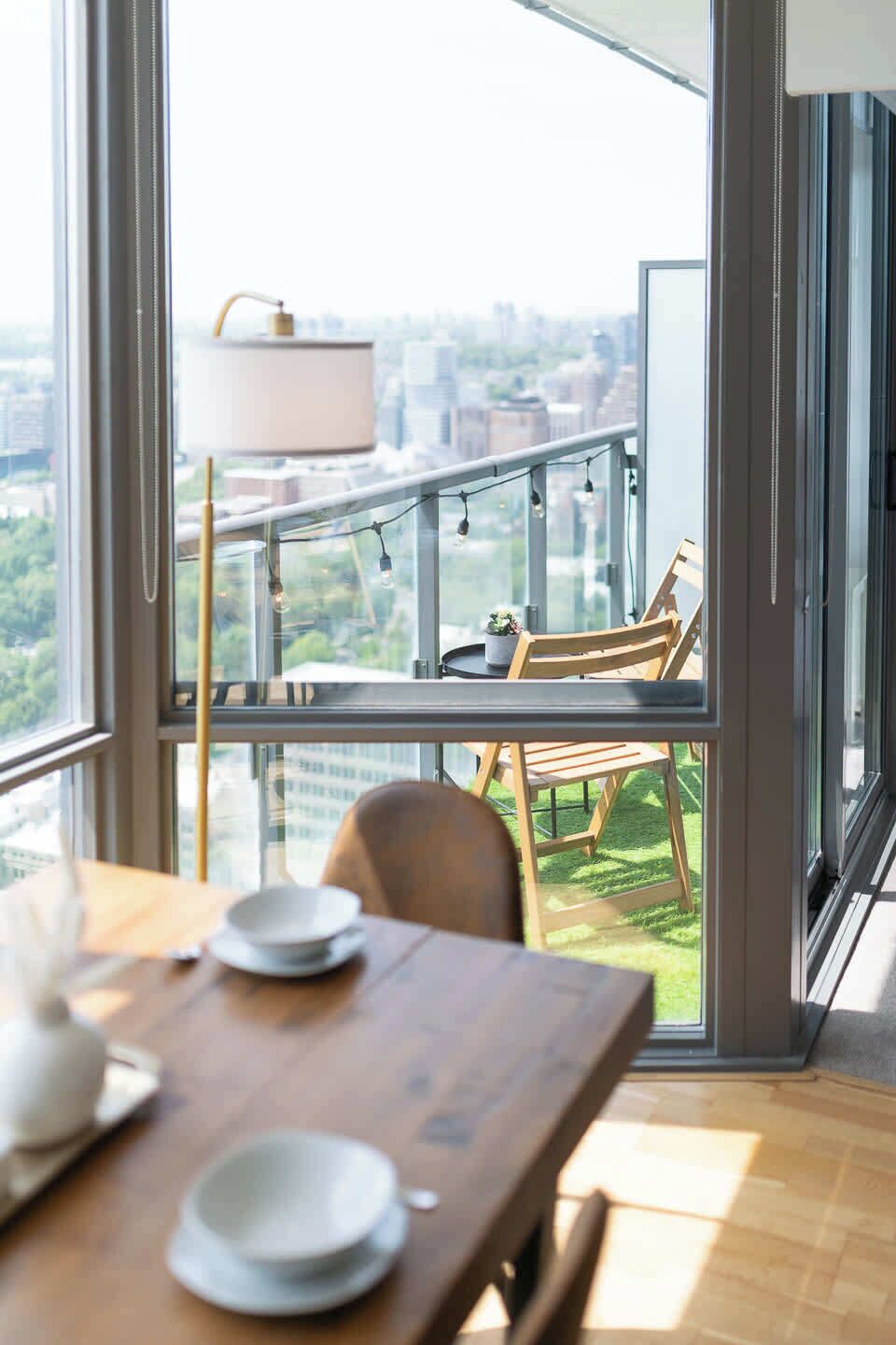 A dining area with a table and chairs, a view of a cityscape through large windows, and a balcony with a railing and string lights.