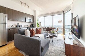A modern living room with a grey sofa, a dining area with a table and chairs, a kitchen with dark wood cabinets, and a large window offering a city view. - Financial District 3BR CN Tower Views! (Toronto)