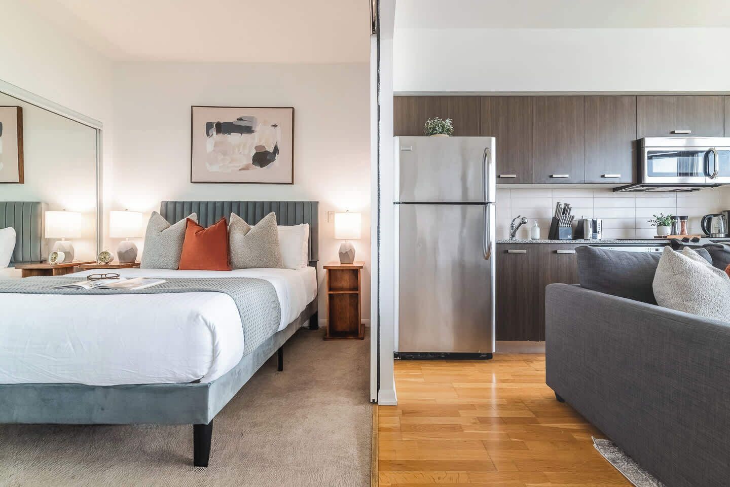 Une chambre d’hôtel moderne comprenant un lit, des tables de chevet, un canapé gris, un coin cuisine avec un réfrigérateur et un four à micro-ondes, et un tableau accroché au mur.