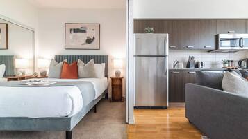 Une chambre d’hôtel moderne comprenant un lit, des tables de chevet, un canapé gris, un coin cuisine avec un réfrigérateur et un four à micro-ondes, et un tableau accroché au mur.