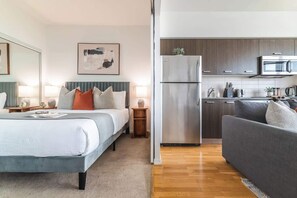 A modern hotel room with a bed, bedside tables, a grey sofa, a kitchen area with a refrigerator and microwave, and a painting on the wall.
