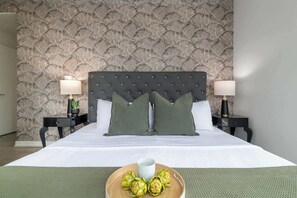 Ein Hotelzimmer mit einer gepolsterten Kopfstütze, zwei Nachttischen mit Lampen, einem Bett mit weißen Bettwäsche und einem Tablett mit Früchten auf dem Bett.