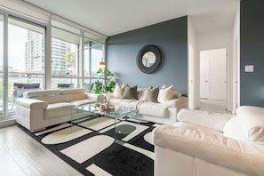 A modern living room with a glass coffee table, two sofas, a large mirror, and a view of a cityscape through large windows. - Upscale 3BR Downtown Suite with Parking! | Parking Available (Toronto)