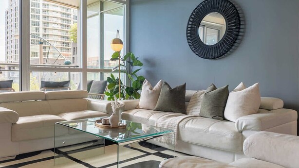 A modern living room with a glass coffee table, a beige sofa, and a large window offering a city view. - Upscale 3BR Downtown Suite with Parking! (Toronto)