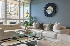 A modern living room with a glass coffee table, a beige sofa, and a large window offering a city view. - Upscale 3BR Downtown Suite with Parking! | Parking Available (Toronto)