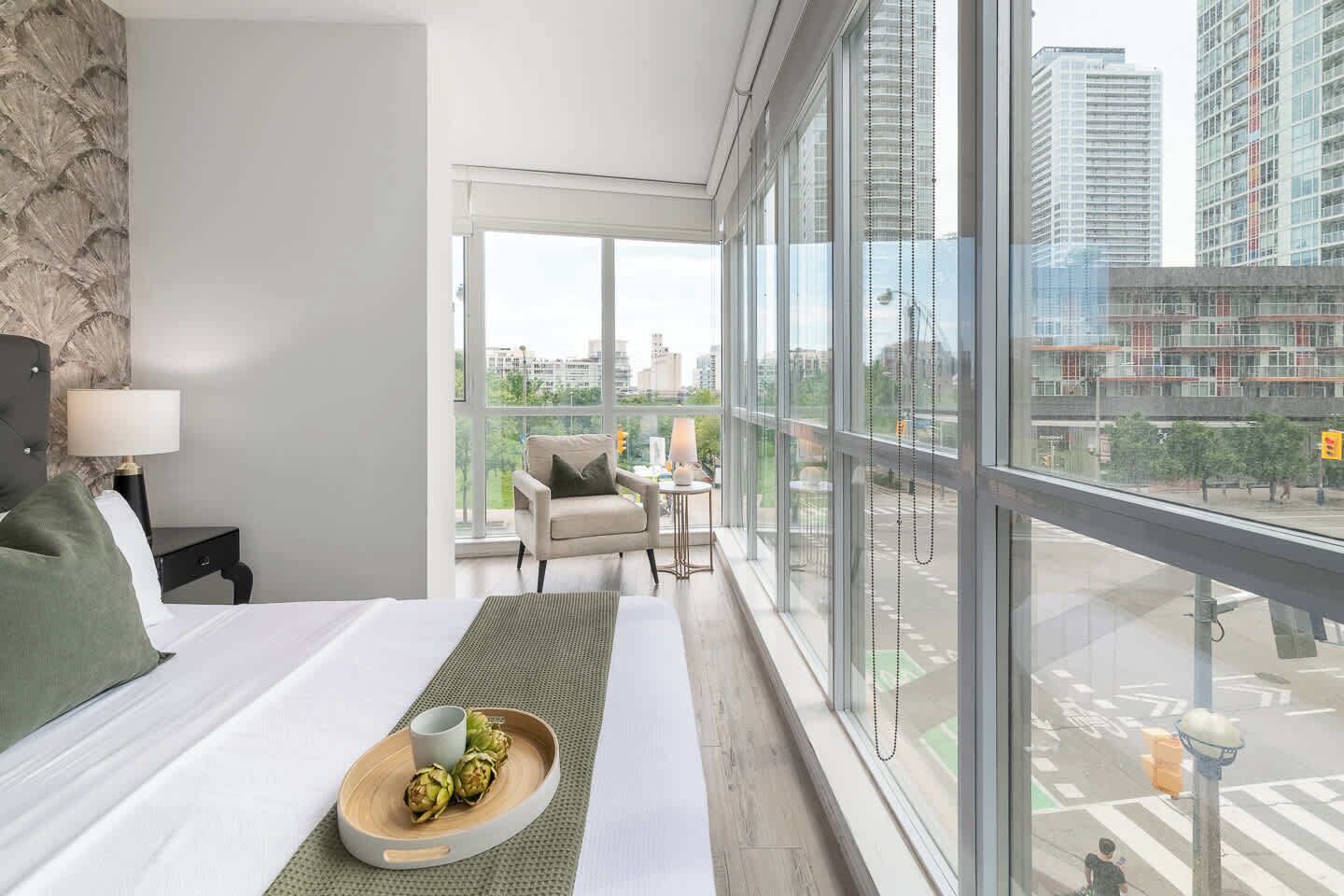 A hotel room with a large window, a bed, a chair, a small table, and a tray with food.