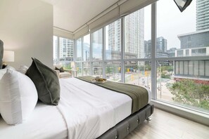 A modern bedroom with a large bed, a view of the city through floor-to-ceiling windows, and a cityscape visible outside. - Upscale 3BR Downtown Suite with Parking! | Parking Available (Toronto)