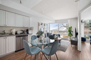 A modern kitchen with white cabinets, a glass dining table, and a view of the city through large windows. - Downtown Condo Walk to CN Tower & Entertainment (Toronto)