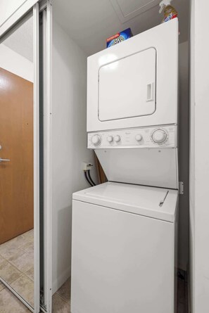 A compact, white laundry room with a front-loading washer and dryer, a small shelf with a bottle, and a door leading to another room.