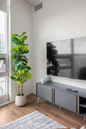 A modern living room with a large flat-screen TV mounted on the wall, a potted plant, a grey console table with a remote control, and a wooden floor.