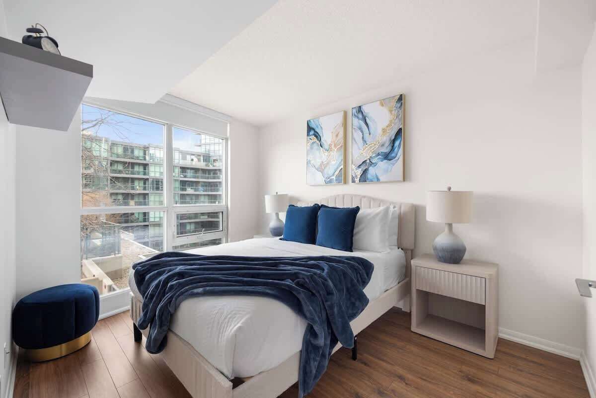 A bedroom with a bed, a nightstand, a lamp, a window, and abstract art on the wall.