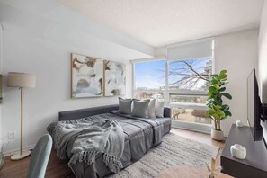 Ein modernes Schlafzimmer mit einem großen Bett, einem Schreibtisch, einem Stuhl und einem Fenster mit Blick auf Gebäude.