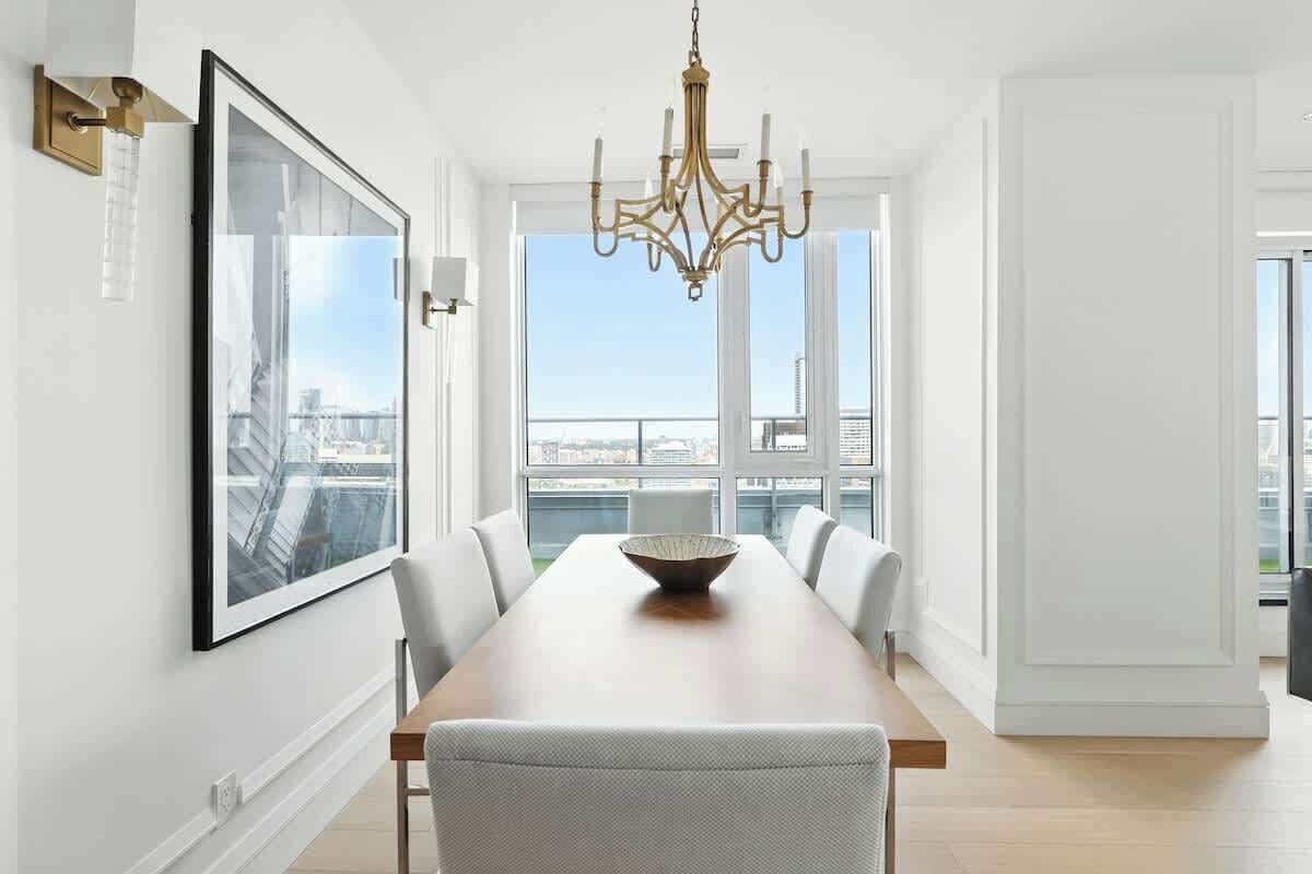 A dining area with a long table, chairs, a chandelier, a framed picture, and a view of the city through large windows.