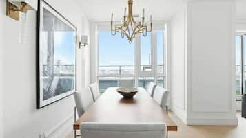 A dining area with a long table, chairs, a chandelier, a framed picture, and a view of the city through large windows.