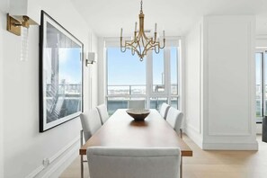 A dining area with a long table, chairs, a chandelier, a framed picture, and a view of the city through large windows. - 2BR Entertainment District Hub | Balcony & Parking | Parking Available (Toronto)