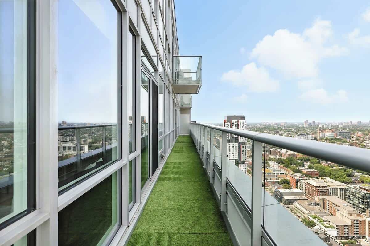 A high-rise building with a narrow balcony featuring a green artificial grass path, large windows, and a cityscape view.