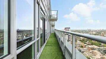 A high-rise building with a narrow balcony featuring a green artificial grass path, large windows, and a cityscape view.