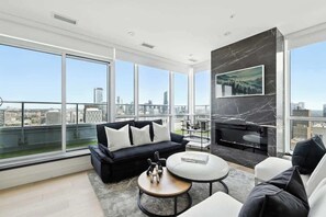 A modern living room with a black sofa, white armchairs, a round coffee table, and a fireplace. Large windows offer a city view. - 2BR Entertainment District Hub | Balcony & Parking | Parking Available (Toronto)