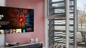 Una sala de estar moderna con un televisor de pantalla plana grande montado en una pared rosa, una mesa de centro de vidrio y un ventanal corredero que da a un área exterior.