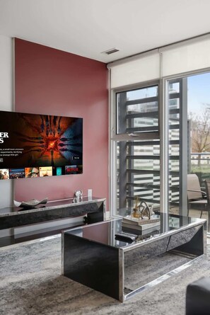 A modern living room with a large flat-screen TV mounted on a pink wall, a glass coffee table, and a sliding glass door leading to an outdoor area.