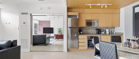 A modern kitchen with stainless steel appliances, a dining area with a round table and chairs, and a living area with a sofa and TV.