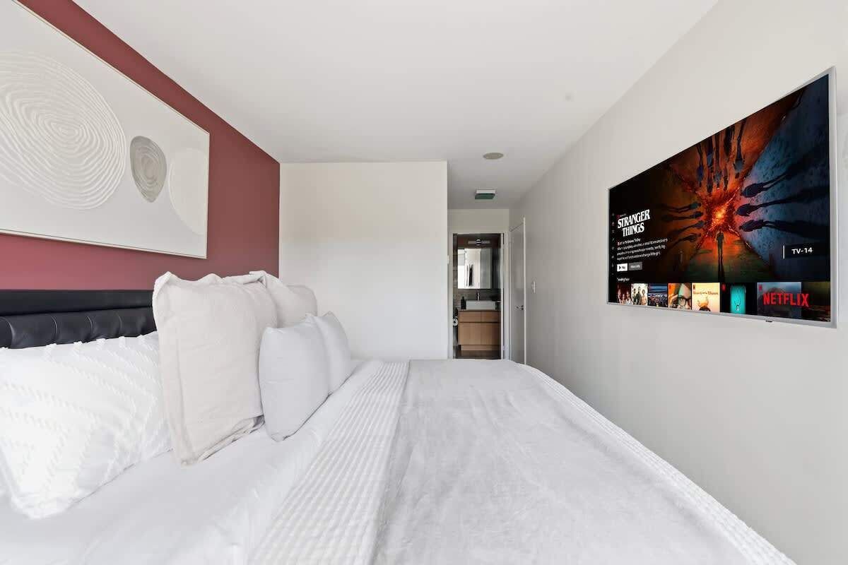 A modern hotel room with a large bed, a mounted TV displaying a movie poster, and a minimalist decor.