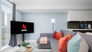 Ein modernes Wohnzimmer mit einem Flachbildfernseher, der das Netflix-Logo zeigt, einem grauen Sofa mit bunten Kissen, einer Stehlampe und einer kleinen Pflanze auf einem Tablett.
