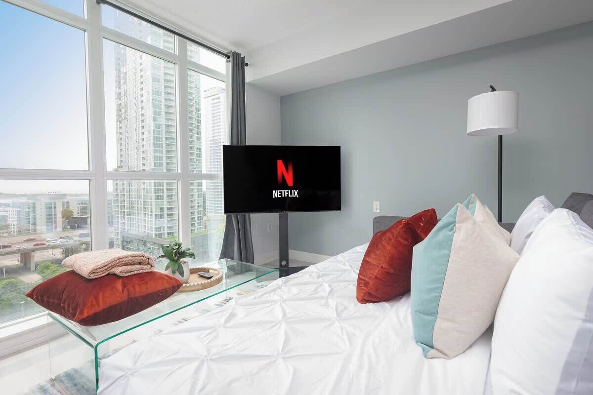 A modern hotel room with a large window, a flat-screen TV displaying the Netflix logo, a glass coffee table, and a bed with multiple pillows.