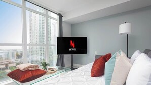 Ein modernes Hotelzimmer mit großem Fenster, einem Flachbildfernseher, der das Netflix-Logo zeigt, einem Glastisch und einem Bett mit vielen Kissen.