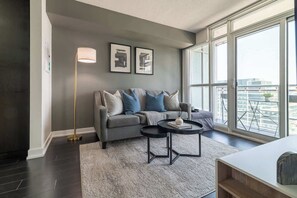 Ein modernes Wohnzimmer mit einem grauen Sofa, einem runden Couchtisch und einer Stehlampe. An den Wänden hängen zwei gerahmte Bilder und ein großes Fenster bietet Blick auf die Stadt.