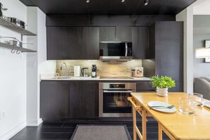A modern kitchen with dark cabinetry, stainless steel appliances, and a wooden dining table with a potted plant. - Modern City Escape 1BR Downtown Toronto (Toronto)