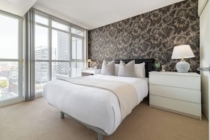 A hotel room with a large bed, a nightstand, a lamp, and a view of the city through large windows. - Modern City Escape 1BR Downtown Toronto (Toronto)