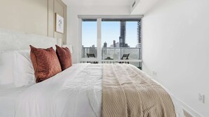 A bedroom with a large bed, a city view through the window, and a modern design. - Modern 1BR Suite Steps to Subway City Views! (Toronto)