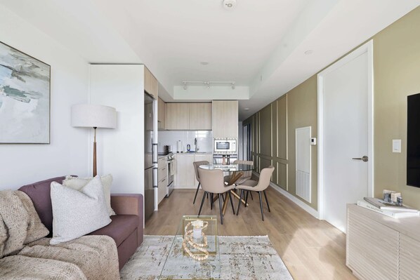 A modern living room with a sofa, a coffee table, and a lamp. There is a dining area with a table and chairs. The kitchen is visible in the background with cabinets and appliances.