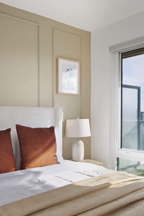 Ein Hotelzimmer mit Bett, Kissen, einem Nachttisch mit Lampe, einem gerahmten Bild an der Wand und einem Fenster mit Aussicht.