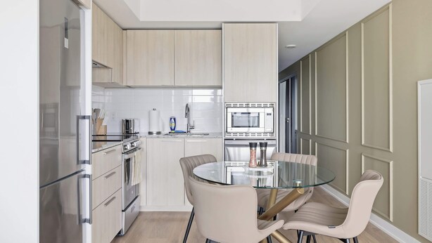 A modern kitchen with stainless steel appliances, a glass dining table, and beige chairs. - Modern 1BR Suite Steps to Subway City Views! (Toronto)