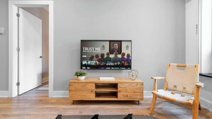 Una sala moderna con un televisor de pantalla plana montado en la pared, un mueble de madera para entretenimiento, un sillón acolchado y una puerta que da a otra habitación.