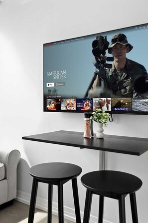 A flat-screen TV mounted on a wall displaying a movie with a military theme, a vase with flowers on a table, and two black stools. - Sleek studio near Eaton Centre in central downtown (Toronto)