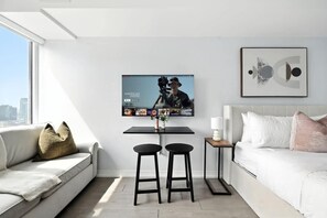 A modern living room with a flat-screen TV mounted on the wall, a small table with two stools, a white sofa with cushions, and a framed abstract artwork above the sofa. - Sleek studio near Eaton Centre in central downtown (Toronto)