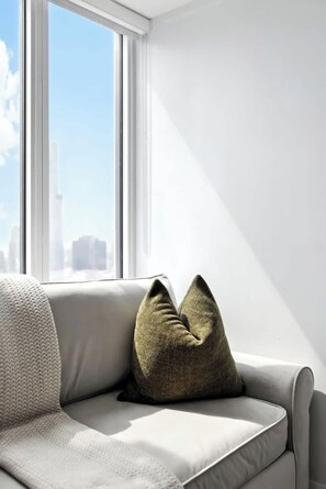 A modern living room with a sofa and two pillows, a large window with a city view, and a white wall. - Sleek studio near Eaton Centre in central downtown (Toronto)