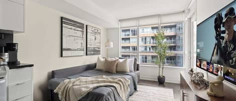 A modern bedroom with a bed, a television, a window with a view of a building, and a potted plant.
