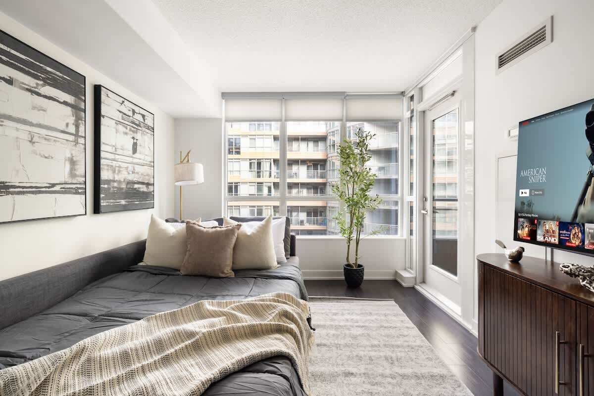 Une chambre moderne avec un grand lit, une plante en pot, un téléviseur et une vue sur un immeuble de grande hauteur.