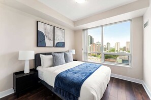 Ein modernes Schlafzimmer mit großem Fenster, einem Bett mit weißer Bettwäsche und einer blauen Decke, zwei Nachttischlampen und abstrakter Kunst an der Wand.