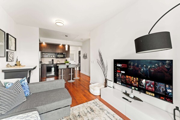 Una sala de estar moderna con un sofá gris, un televisor de pantalla plana montado en la pared y una cocina al fondo.