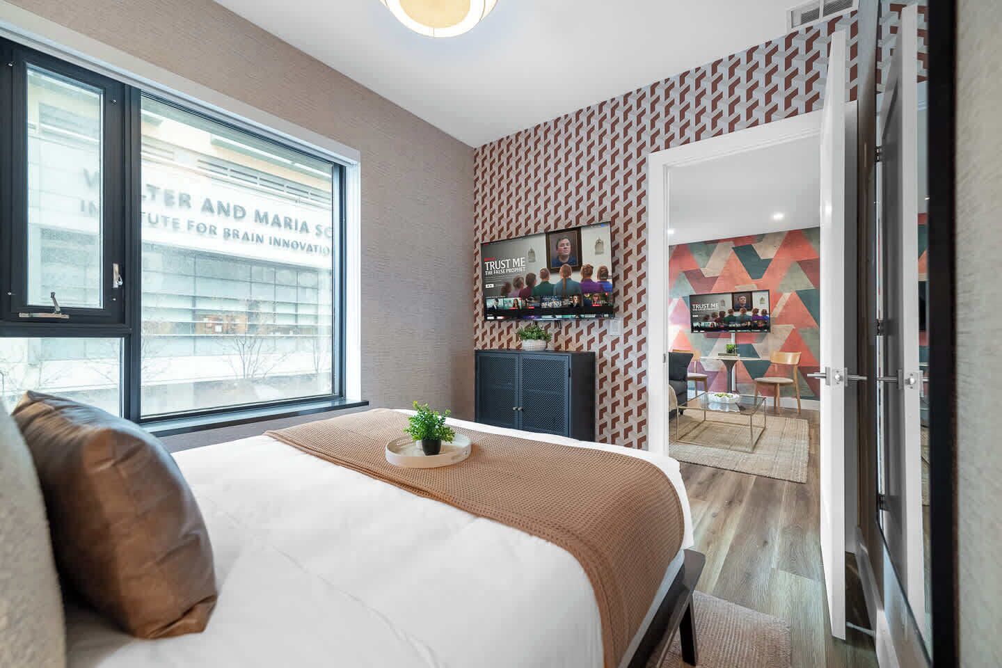 A modern bedroom with a large bed, a window, a desk with a computer, and a patterned wall.
