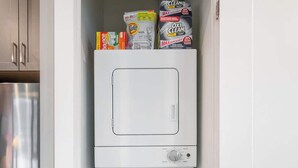 Máquina de lavar compacta e branca, com painel de controle e porta transparente, inserida em um armário estreito com itens de armazenamento acima.