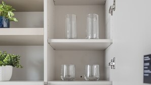 Um banheiro com pia branca, uma torneira, porta-papel higiênico e prateleiras com garrafas de vidro transparente e uma planta em vaso.