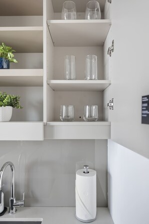 Ein Badezimmer mit einem weißen Waschbecken, einem Wasserhahn, einem Toilettenpapierhalter und Regalen mit klaren Glasflaschen und einer Topfpflanze.